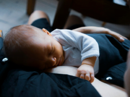 A baby sleeps in a person’s lap, showing how they crave the mother’s closeness even as they rest, contributing to sleep deprivation in new moms
